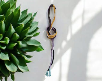 May include: A multi-colored cord with a knot tied in the middle. The cord is made of yellow, purple, black, and white thread. The ends of the cord are tied with a blue tassel.