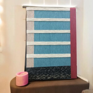 May include: A blue and white striped wall hanging with a red border. The hanging is made of woven yarn and has a black wood frame. A pink candle sits on a brown table in front of the hanging.