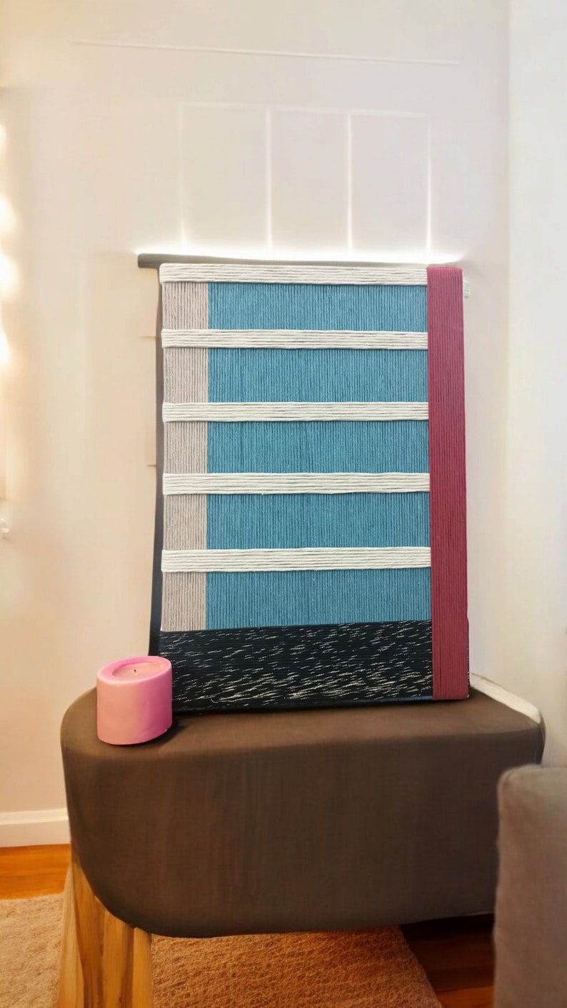 May include: A blue and white striped wall hanging with a red border. The hanging is made of woven yarn and has a black wood frame. A pink candle sits on a brown table in front of the hanging.