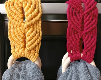Puede incluir: Dos colgadores de toallas de macramé, uno amarillo y otro rojo, colgados de una puerta de horno de plata. Cada colgador tiene un anillo de madera en la parte inferior y una toalla gris cuelga de cada anillo.