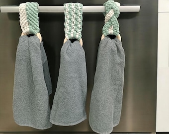 Puede incluir: Tres toallas de cocina grises colgadas en un toallero plateado. Cada toalla tiene un lazo de crochet verde y blanco diferente, unido a un anillo de madera.