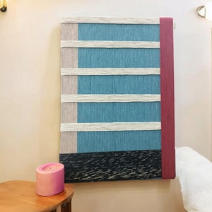 May include: A wall hanging made of woven yarn in shades of blue, white, and pink. The hanging has a black and white striped base and a red border.