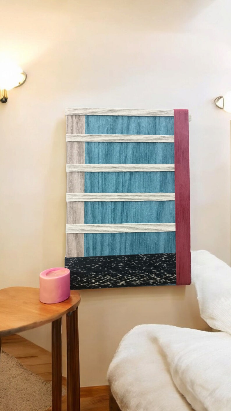 May include: A wall hanging made of woven yarn in shades of blue, white, and pink. The hanging has a black and white striped base and a red border.