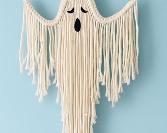 May include: A handmade macrame ghost wall hanging. The off-white ghost features black eyes and a mouth, with long fringe detailing the bottom. A decorative Halloween accent, perfect for adding a spooky touch to any room.