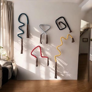 May include: Five colorful, abstract wall hangings. The shapes include a wavy blue line, a gray figure eight, a black square, a red curved line, and a yellow zig-zag. Each piece has a brown tassel at the bottom. The art is mounted on a white wall.