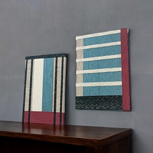 May include: Two abstract wall art pieces with a woven texture. The piece on the left is a vertical rectangle with black, white, and blue stripes and a red band at the bottom. The piece on the right is a square with a black, white, and blue striped pattern and a red band on the right side. Both pieces are hanging on a gray wall above a dark brown wooden bench.