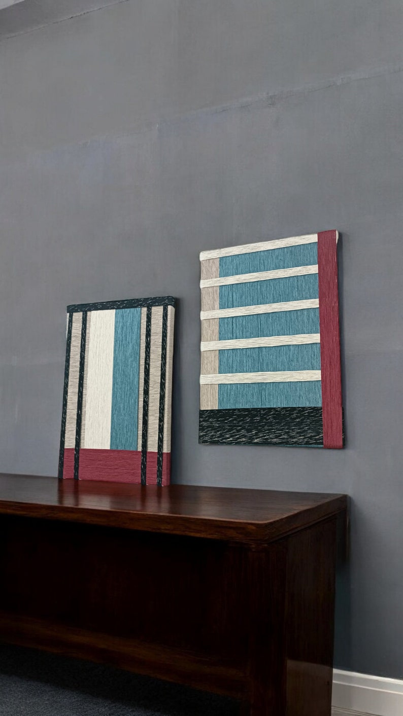 May include: Two abstract wall art pieces with a woven texture. The piece on the left is a vertical rectangle with black, white, and blue stripes and a red band at the bottom. The piece on the right is a square with a black, white, and blue striped pattern and a red band on the right side. Both pieces are hanging on a gray wall above a dark brown wooden bench.