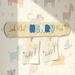 May include: Wooden sign with the text "Look What HENRY Made" in a child's room. The name "HENRY" is in blue letters. Two drawings are clipped to the sign. The wallpaper has cartoon animals and stars.