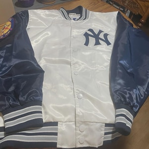 May include: A white and navy blue satin baseball jacket. The jacket displays the New York Yankees logo on the chest and a patch on the left sleeve. The cuffs and hem have striped detailing.