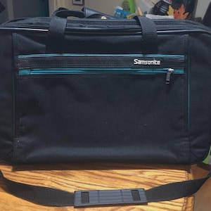 May include: Black Samsonite carry-on bag with a teal zipper accent and the brand name in white. The bag has a top handle, a shoulder strap, and a front zippered pocket. The bag is rectangular and appears to be made of a durable material.