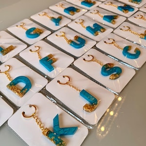 Może przedstawiać: Kolekcja breloków z alfabetem. Każda litera ma żywy turkusowy kolor ze złotymi akcentami, przymocowana do złotego łańcuszka i breloka. Breloki są prezentowane na pojedynczych białych kartach.