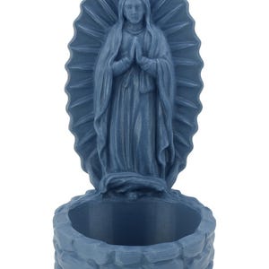 May include: A blue statue of the Virgin Mary with hands clasped in prayer. The statue is set against a radiating halo and sits atop a small, stone-like basin, suitable for a small plant or candle.
