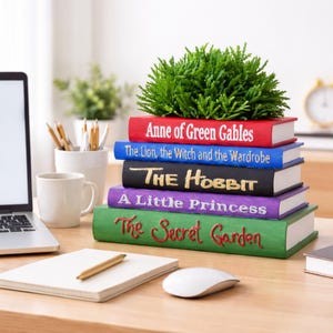 May include: A stack of colorful book-shaped planters with titles including "Anne of Green Gables" and "The Hobbit." Each planter is a different color and holds a small green plant. The planters are on a wooden desk with a laptop and notepad.