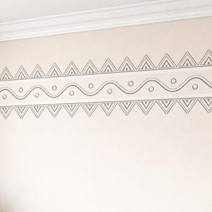 May include: A decorative wall border featuring a repeating geometric pattern in gray. The design includes triangles, wavy lines, and circles, creating a symmetrical and artistic look. The border is applied on a light beige wall.