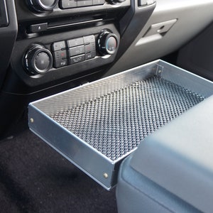 May include: A silver metal tray with a mesh bottom is fitted in the centre console of a car. The tray is designed to hold small items and is located between the driver and passenger seats.
