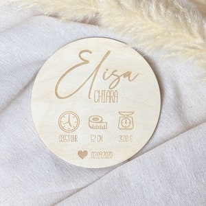 May include: A wooden round plaque with the name "Elisa Chiara" engraved on it. The plaque also includes icons for time, length, and weight, with the corresponding measurements: 03:51 Uhr, 52 cm, and 3500 g. The date "02.09.2020" is also engraved on the plaque.
