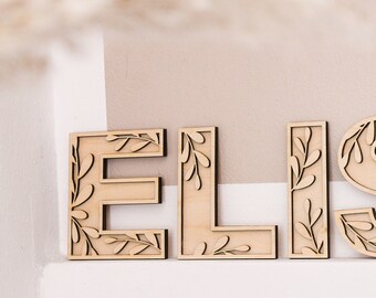 Op de afbeelding: Houten letters die de naam "ELISA" spellen met een bladerontwerp. De letters zijn lichtbruin en hebben een natuurlijke houten afwerking.