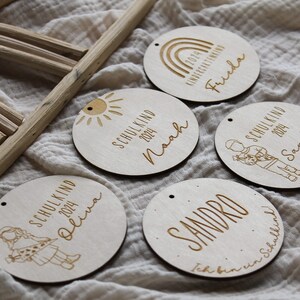 May include: Wooden circular ornaments with engraved text and illustrations. Each ornament features a name, the year 2024, and either "Schulkind" or "Kindergartenkind". Designs include a sun, rainbow, and children.