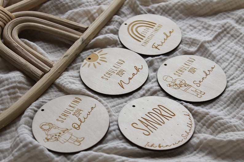 May include: Wooden circular ornaments with engraved text and illustrations. Each ornament features a name, the year 2024, and either "Schulkind" or "Kindergartenkind". Designs include a sun, rainbow, and children.