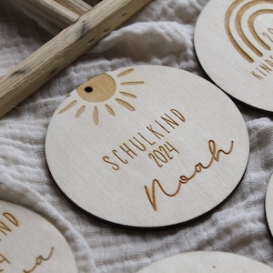 May include: Wooden round ornaments with engraved text and designs. One ornament features a sun design and the text "SCHULKIND 2024 Noah". Other ornaments have rainbow and child designs. The ornaments are light brown.