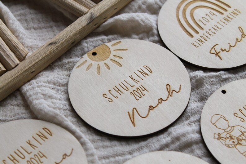 May include: Wooden round ornaments with engraved text and designs. One ornament features a sun design and the text "SCHULKIND 2024 Noah". Other ornaments have rainbow and child designs. The ornaments are light brown.