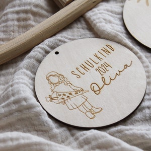 May include: Wooden round ornament with engraved text and illustration. The ornament reads "SCHULKIND 2024 Olivia" with a drawing of a child holding a school cone. The ornament has a small hole at the top for hanging.