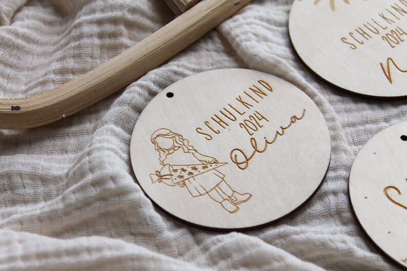 May include: Wooden round ornament with engraved text and illustration. The ornament reads "SCHULKIND 2024 Olivia" with a drawing of a child holding a school cone. The ornament has a small hole at the top for hanging.