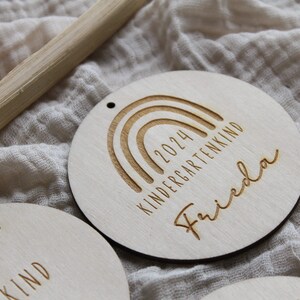May include: Wooden round ornament with a rainbow design, the text "2024 KINDERGARTENKIND" and the name "Frieda" engraved. The ornament has a small hole at the top for hanging. Other similar ornaments are visible.