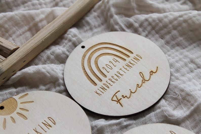 May include: Wooden round ornament with a rainbow design, the text "2024 KINDERGARTENKIND" and the name "Frieda" engraved. The ornament has a small hole at the top for hanging. Other similar ornaments are visible.
