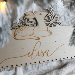 May include: A wooden sign with a laser-cut design of a deer and snowflakes. The name "elisa" is written in cursive below the deer. The sign has a row of holes along the bottom edge.