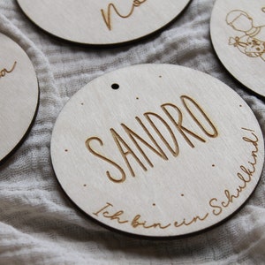 May include: Wooden circular ornaments with engraved names and the year 2024. One ornament says "Sandro" with "Ich bin ein Schulkind!" below. Other ornaments have names like "Olivia" and "Sasha".