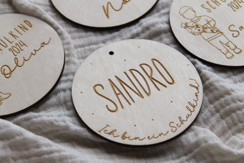 May include: Wooden circular ornaments with engraved names and the year 2024. One ornament says "Sandro" with "Ich bin ein Schulkind!" below. Other ornaments have names like "Olivia" and "Sasha".