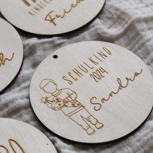 May include: Wooden round ornaments with engraved text and illustrations. One ornament features a child carrying a school cone, the text "SCHULKIND 2024 Sandra". Other ornaments have names and "SCHULKIND 2024" engraved.