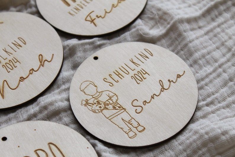 May include: Wooden round ornaments with engraved text and illustrations. One ornament features a child carrying a school cone, the text "SCHULKIND 2024 Sandra". Other ornaments have names and "SCHULKIND 2024" engraved.