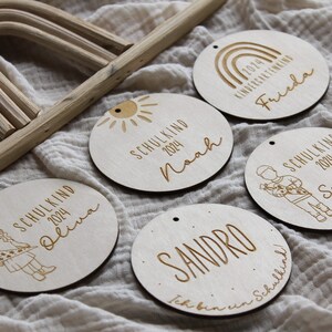 May include: A collection of six wooden round ornaments with engraved text and illustrations. Each ornament features a name, the year 2024, and the word "SCHULKIND" or "KINDERGARTENKING". The ornaments are light brown with a small hole for hanging.