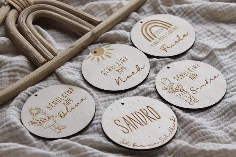 May include: A collection of six wooden round ornaments with engraved text and illustrations. Each ornament features a name, the year 2024, and the word "SCHULKIND" or "KINDERGARTENKING". The ornaments are light brown with a small hole for hanging.