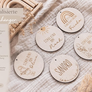 May include: Wooden round ornaments with engraved names and the year 2024. The ornaments feature illustrations of a rainbow, sun, and children. Text on the ornaments includes "Kindergartenkind", "Schulkind", and names like "Frieda", "Noah", "Sandra", "Oliva", and "Sandro".