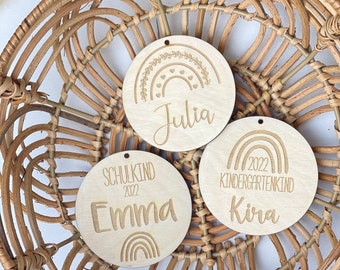 Könnte beinhalten: Drei Holzornamente mit Regenbogen-Designs und personalisierten Namen. Die Ornamente sagen "Julia", "Emma - Schulkind 2022" und "Kira - Kindergartenkind 2022".