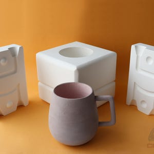 May include: Three white ceramic molds for making a mug. The molds are stacked on top of each other. A purple mug is sitting in front of the molds.