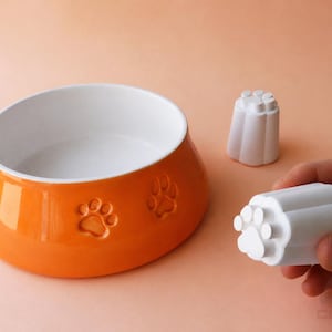 Può includere: Ciotola arancione per animali domestici con interno bianco e dettagli di impronte di zampe. Include due stampi bianchi a forma di zampa. La ciotola è di colore arancione lucido.