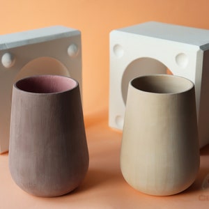 May include: Two ceramic mugs, one brown and one beige, are displayed next to their white plaster molds. The molds have a circular opening in the center.