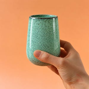 May include: A cylindrical ceramic tumbler featuring a speckled mint green glaze with a dark brown rim. The vessel has a smooth, rounded base and a straight-sided body, showcasing a reactive finish with fine dark flecks throughout the surface.