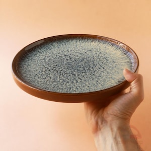 May include: A brown ceramic plate with a speckled blue and white interior. The plate is held by a hand against a peach-coloured background. The plate has a glossy finish and a slightly raised rim.