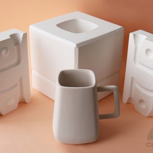May include: White ceramic mug with a square shape and a handle. The mug is surrounded by three white ceramic moulds.