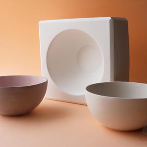 Pode incluir: Duas tigelas de cerâmica, uma rosa e outra branca, sobre uma superfície laranja claro. Um molde quadrado branco com uma indentação redonda fica atrás das tigelas.