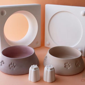 Peut inclure: Deux moules en céramique blanche pour faire des bols pour animaux de compagnie. Les moules sont carrés et ont une ouverture ronde au centre. Deux bols pour animaux de compagnie finis sont présentés devant les moules. Un bol est rose et l'autre est beige. Les deux bols ont des empreintes de pattes sur le fond.