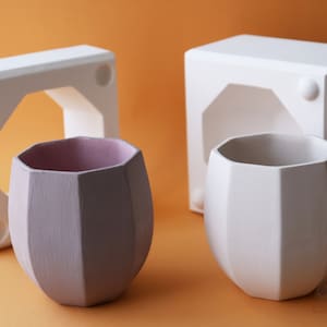 Op de afbeelding: Twee witte keramische mallen voor het maken van geometrische plantenbakken. Een mal is een vierkant met een rechthoekige opening. De andere mal is een ronde plantenbak met een geometrisch ontwerp. Een paarse plantenbak staat naast de mallen.