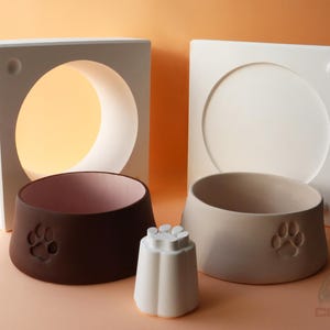 以下が含まれることがあります： 茶色とベージュのセラミック製ペットボウルのセット。それぞれに足跡のデザインが施されています。ボウルは白い幾何学的な構造と組み合わされています。小さな白い花型のオブジェクトも存在します。