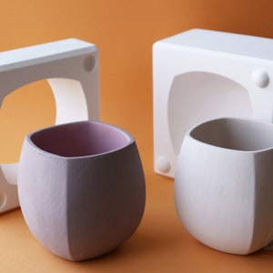 May include: Two ceramic planters, one white and one light purple, are shown next to their white plaster molds. The planters have a rounded, slightly squared shape.