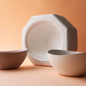 May include: A studio shot features three ceramic pieces: two bowls and a large, geometric, white form. The bowls are in shades of brown and beige. The background is a warm, peachy tone, creating a minimalist aesthetic.